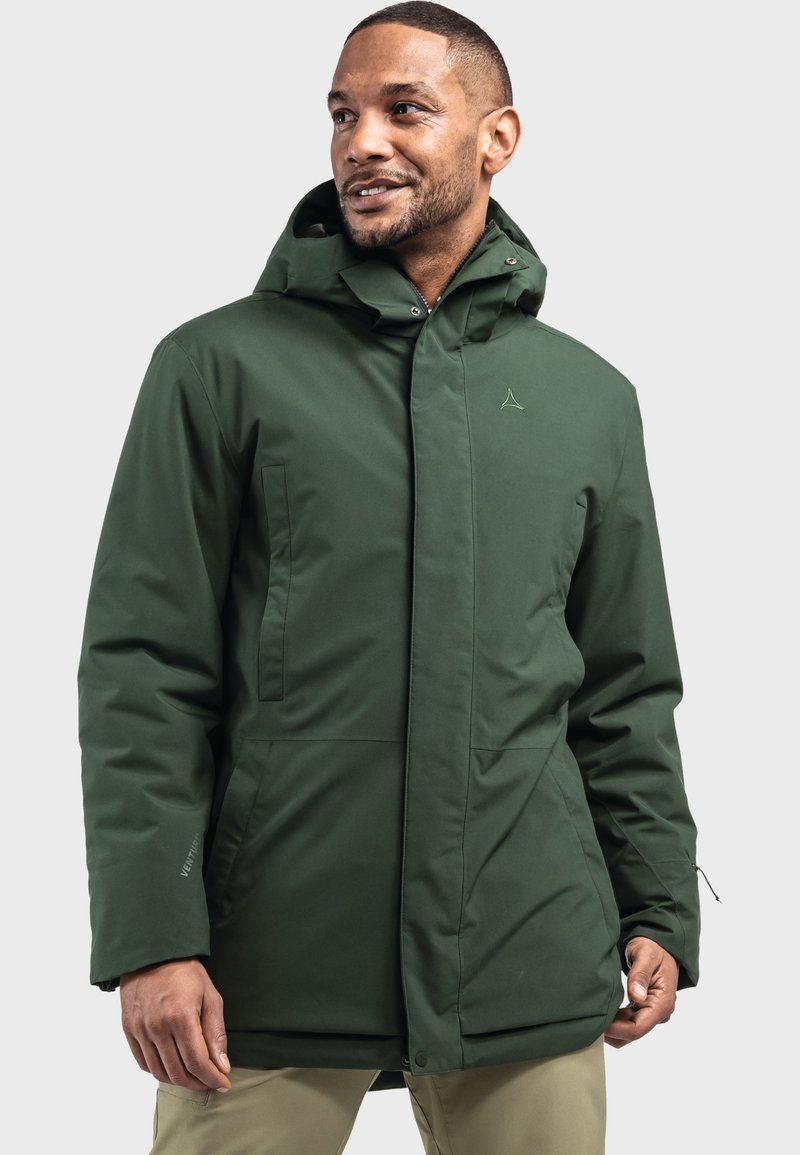 Mann trägt eine dunkelgrüne Kapuzen-Winterjacke mit Vordertaschen und beigen Hosen, steht vor einem schlichten hellen Hintergrund.