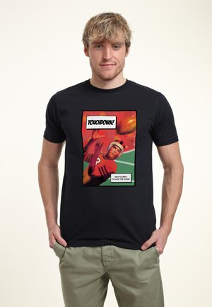 Mann trägt schwarzes T-Shirt mit farbenfrohem Comic-Stil-Aufdruck eines Footballspielers, der den Ball fängt, Text: "Touchdown! Er hat zum Gewinn des Spiels gepunktet."