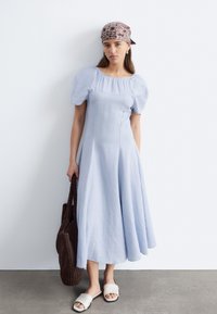 Femme portant une robe midi bleu clair avec des manches bouffantes, un bandana à motif, des sandales blanches à enfiler, et tenant un grand sac fourre-tout tricoté marron foncé.