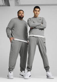 To modeller bærer matchende grå sweatshirts og bukser lavet af blødt stof, der har et lille sort logo på brystet og taljen. Hvide sneakers fuldender looket.
