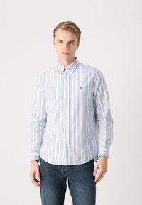 Blau-weiß gestreiftes Button-down-Hemd mit langen Ärmeln, ausgestattet mit einem kleinen Logo auf der Brust und einem abgerundeten Saum. Getragen mit dunklen Jeans.