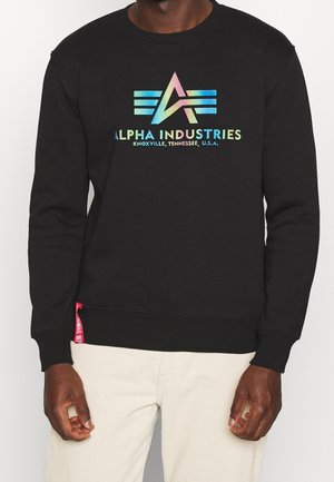 Mężczyzna ubrany w czarną bluzę z kolorowym logo Alpha Industries i napisem "Knoxville, Tennessee, USA", zestawioną z jasnobeżowymi spodniami.