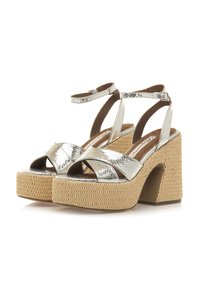Sandalias de plataforma metálica plateada con tiras entrecruzadas y un tacón texturizado similar a la paja. El tacón presenta una forma recortada.