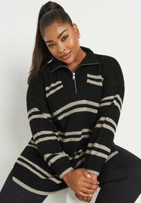 Pull noir en tricot avec col zippé, présentant des rayures horizontales beiges, une texture côtelée et une coupe oversize, porté avec un pantalon foncé.