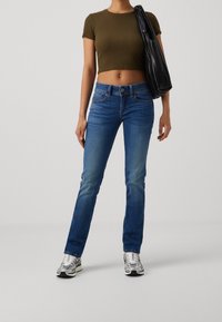 Blå denimjeans med rak passform, i kombination med en kort olivgrön topp och silverfärgade sneakers. Har knappstängning fram och fickor.