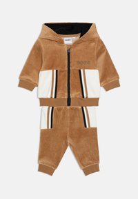 Tuta in velluto tan e bianco con cappuccio zip, dettagli neri e strisce orizzontali. Presenta tasche e polsini a coste.