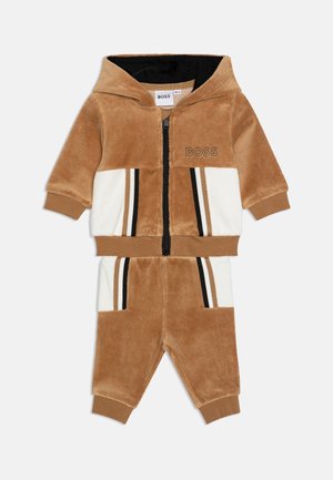 Tannenkleurig en wit velours trainingspak met een rits hoodie, zwarte accenten en horizontale strepen. Bevat zakken en ribgebreide cuffs.