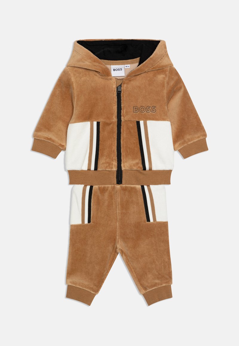 Tuta in velluto tan e bianco con cappuccio zip, dettagli neri e strisce orizzontali. Presenta tasche e polsini a coste.