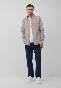 Man in een wit gestreept overhemden met knopen over een wit T-shirt, navy broek en witte sneakers, staande tegen een effen achtergrond.