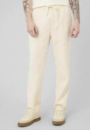 Personne portant un pantalon à cordon de serrage écru et des baskets assorties, debout avec des tatouages visibles sur les bras devant un fond uni.