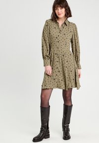Robe vert olive avec imprimé floral, col boutonné, manches longues bouffantes et jupe évasée ; associée à des bottes noires jusqu'aux genoux.