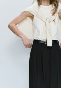 Weißes ärmelloses Top, kombiniert mit hochtaillierten schwarzen Hosen. Ein helles Cremefarbenes Sweatshirt, über die Schultern gelegt, schwarzer Gürtel mit rechteckiger Schnalle.