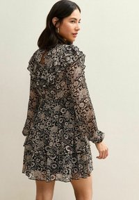 Zwarte bloemenjurk met doorschijnende stof, gerimpelde halslijn en mouwen, getailleerde taille en gelaagde rokdetails. Het patroon bevat lichte bloemdecoraties.