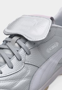 Κοντινό πλάνο ενός ασημί-γκρι δερμάτινου αθλητικού παπουτσιού με γκρι κορδόνια, διπλωμένη γλώσσα και λογότυπο "KING" στην πλευρική επιφάνεια.