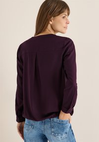 Langærmet bluse i dyb lilla stof, med en afslappet pasform, bagplise detaljer og knaplukning ved håndleddene, parret med lyse denim.