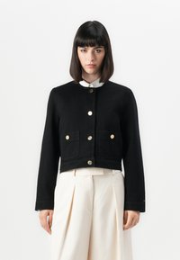 Veste noire courte en denim avec des manches longues, deux poches avant et des fermetures à boutons dorés. Portée par-dessus une chemise blanche à col.
