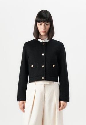 Black cropped jacket made from denim with long sleeves, two front pockets, and gold button fastenings. Styled over a white collared shirt.