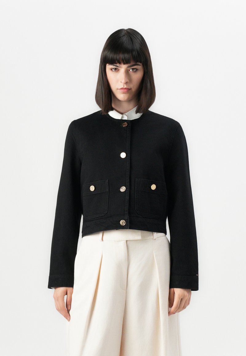 Veste noire courte en denim avec des manches longues, deux poches avant et des fermetures à boutons dorés. Portée par-dessus une chemise blanche à col.