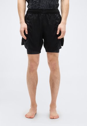 Homme debout pieds nus portant un short de sport noir avec poches et une chemise noire à motifs, vu de la taille vers le bas sur un fond uni.