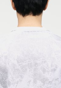 Άτομο με κοντά μαύρα μαλλιά, που φαίνεται από πίσω, φοράει ένα λευκό πουκάμισο με ανοιχτό γκρι αφηρημένο μοτίβο.