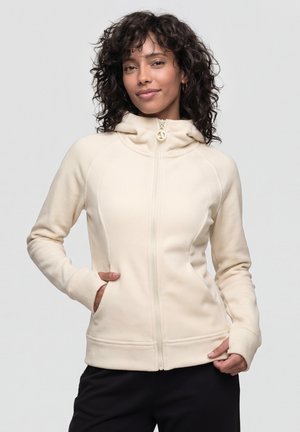 Frau mit lockigem Haar, die einen cremefarbenen Hoodie mit Reißverschluss und Daumenlöchern trägt, steht vor einem einfarbigen grauen Hintergrund.