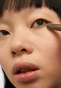 Nærbilde av et ansikt som påfører eyeliner med en mørk blyant. Merkbare trekk inkluderer lys hud, brune øyne og lett parted lepper.