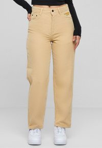 Pantaloni in corduroy beige con taglio dritto, dotati di tasche frontali e un logo ricamato giallo vicino alla vita; abbinati a sneakers bianche.