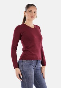 Maglione lavorato a maglia bordeaux con scollo a V, maniche lunghe e silhouette aderente. Tessuto testurizzato con dettagli in costina sull'orlo. Abbinato a dei jeans.