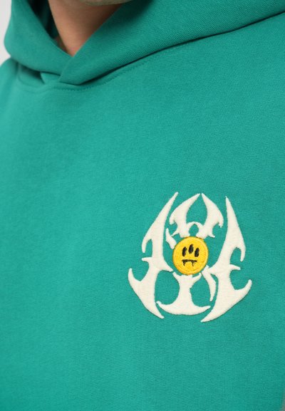 Felpa turchese con un grande logo ricamato bianco che presenta artigli stilizzati e una faccina sorridente con espressione triste. Materiale in cotone misto.