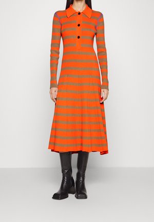 Femme portant une robe midi à manches longues rayée orange et marron avec des boutons noirs, accompagnée de bottes noires à talons montant jusqu'aux genoux.