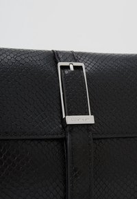 Černá texturovaná kožená klopa tašky se stříbrnou obdélníkovou sponou s logem Calvin Klein.