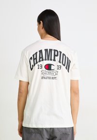 Weißes Baumwoll-T-Shirt mit großem schwarzen "CHAMPION"-Druck auf dem Rücken, mit einem roten und schwarzen Logodetail und dem Text "ATHLETICS DEPT." darunter.