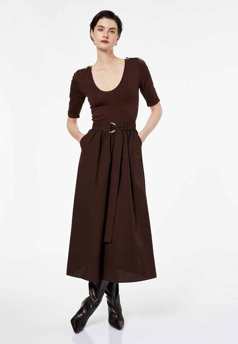 Mujer con un vestido midi marrón oscuro, ceñido con cinturón y mangas tres cuartos, con botones dorados, vistiendo botas negras de punta, de pie con las manos en los bolsillos.