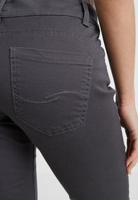 Jeans en denim gris avec une poche arrière ornée de délicates coutures. Le tissu a une texture lisse et est ajusté à la taille.