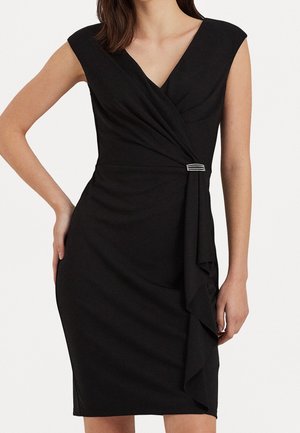 Femme portant une robe noire sans manches longueur genoux avec un col en V et un détail drapé sur le côté fixé par une petite broche rectangulaire.