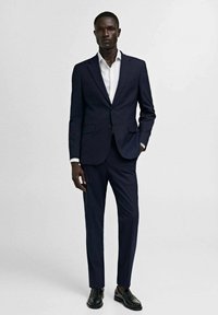 Dunkelblauer maßgeschneiderter Anzug, Ein-Knopf-Blazer, eng geschnittene Hose, weißes Hemd, schwarze Lederschuhe, glattes Material, minimalistisches Design.