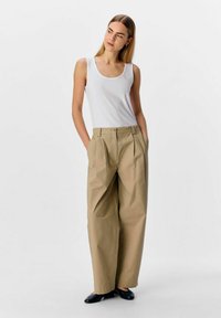 Pantalon large beige avec des plis, en tissu lisse, associé à un débardeur blanc ajusté. Le modèle se tient avec les mains dans les poches.