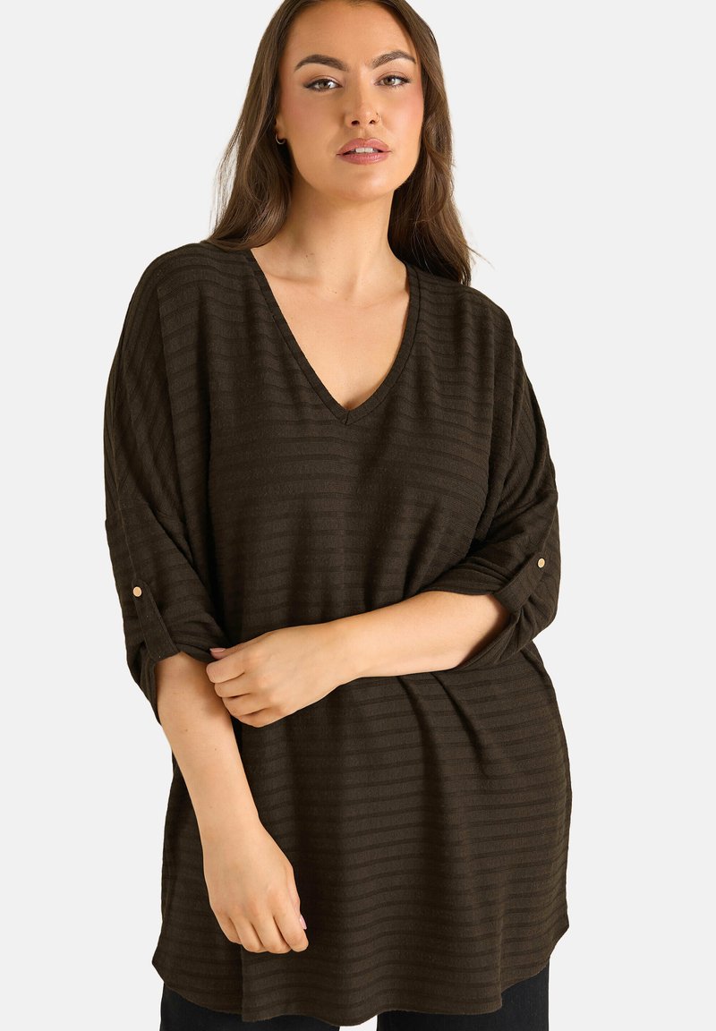 Donna con capelli castani lunghi che indossa una maglia marrone scuro a scollo a V, ampia, con maniche arrotolate, su sfondo neutro.