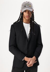 Sort blazer med en skræddersyet pasform, enkeltknappet design, parret med en grå kasket med farverig broderi og lapper.