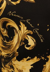 Musta kangas, jossa kultaisia koristeellisia barokkityylisiä kiemuroita ja kultaisella kursiivikirjoituksella teksti "Versace Jeans Couture".