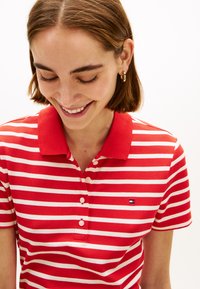 Νεαρή γυναίκα που χαμογελά και κοιτάζει προς τα κάτω, φοράει κοντομάνικη πόλο μπλούζα με ριγέ κόκκινο και λευκό, έχει κοντά καστανά μαλλιά και χρυσούς κρίκους στα αυτιά.