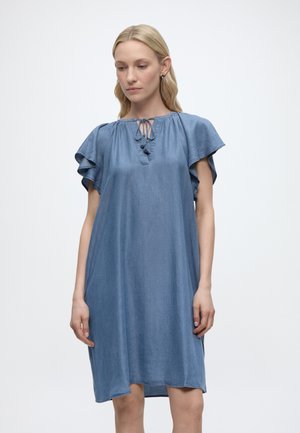 Kobieta ubrana w luźną niebieską sukienkę z krótkimi, zwiewnymi rękawami i wiązanym dekoltem typu keyhole, stojąca na jednolitym białym tle.