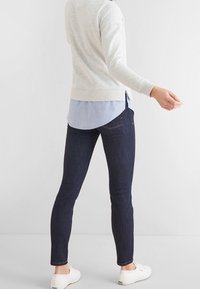 Pull gris clair superposé à une chemise boutonnée bleue, associé à un jean skinny foncé et des baskets blanches, vu de dos.