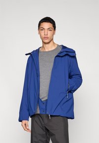 Blå vattentät jacka med huva, med frontdragkedja och fickor. Bärs över en grå skjorta, har en avslappnad passform och slät textur.