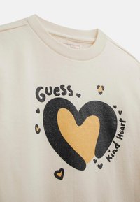 Cremefarvet t-shirt med rund halsudskæring, der har et stort, sort og guld hjertegrafik, omgivet af små hjerter og tekst.