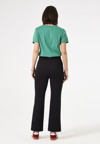 Groene t-shirt met korte mouwen, zwarte strakke broek en rode plateau-sandalen met witte accenten, gezien vanuit de rug tegen een witte achtergrond.