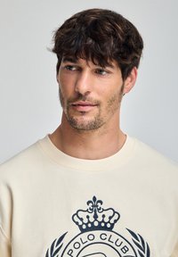 Felpa color crema con un grande logo blu navy che presenta una corona, una laura e del testo. Tessuto morbido e scollo rotondo, con una vestibilità casual.