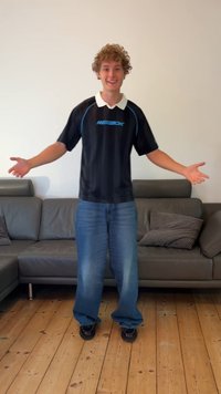 Camisola atlética preta com detalhes azuis, colarinho branco, e jeans azuis largos. De pé sobre um piso de madeira ao lado de um sofá cinza com almofadas.