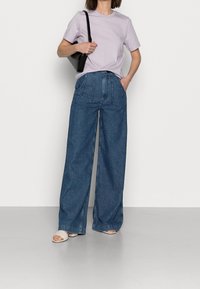 Helles Lavendel-T-Shirt, locker geschnitten, kombiniert mit hochgeschnittenen, weiten blauen Jeans. Schwarze Umhängetasche und hellfarbene Sandalen.