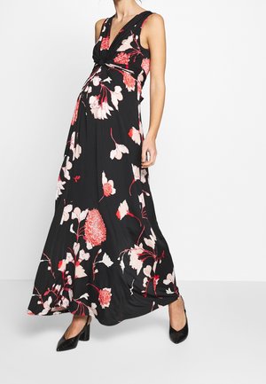 Kobieta w czarnej, bezrękawnikowej sukni maxi w czerwono-biały kwiatowy wzór i czarnych szpilkach, stojąca na białym tle.